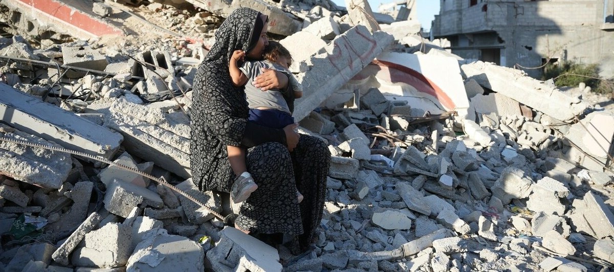 Una donna è seduta tra le macerie di edifici distrutti, tenendo un bambino piccolo tra le braccia. La scena mostra devastazione e perdita a Gaza.